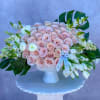 Low compote of blush roses with white tulips and large tropical leaves in a white pedestal vase