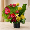 Tropical flower arrangement with red anthuriums, yellow blooms, and roses in a black vase