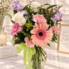 Mixed bouquet of pink, white, and lavender flowers in a glass vase with a green ribbon.