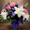 Pink roses, white lilies, and purple daisies in a glass vase with a purple ribbon