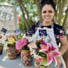 Woman holding colorful flower arrangements in painted ceramic vases outdoors.