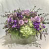 Low pedestal arrangement of lavender roses, purple blooms, and green hydrangeas in a glass compote vase