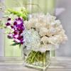 White roses and hydrangeas in a clear cube vase with purple orchids and green Bells of Ireland in the background.