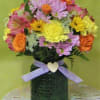 Mixed bouquet of pink, yellow, and orange flowers in a dark vase with a ribbon