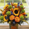 Autumn bouquet with sunflowers, orange roses, green mums, and blue accents in a pumpkin-decorated vase.