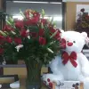 Red roses and lilies in a glass vase beside a white teddy bear with a red bow