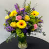 Mixed bouquet of yellow gerbera daisies, pink and yellow roses, and purple flowers in a glass vase