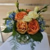 Compact arrangement of peach and cream roses with blue hydrangeas in a clear glass vase with feathery greenery.