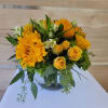 Low arrangement of yellow roses and gerbera daisies in a clear glass vase with green accents