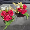Pair of red rose and white accent flower wrist corsages with gold ribbon and fern accents on a reflective black surface.