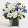 White lilies, roses, and daisies with blue flowers in a square glass vase