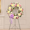 Round floral wreath of white, pink, and lavender flowers on a stand with a purple bow