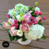 Compact arrangement of pink roses and spray roses with white and green hydrangeas in a white vase
