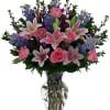 Pink lilies and roses with blue flowers in a clear glass vase