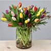 Mixed-color tulip arrangement in a clear glass cylinder vase with visible stems and stones at the base