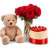 Red roses in a glass vase beside a teddy bear and strawberry cake