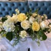 Low arrangement of yellow and cream roses with white hydrangeas on a set table.