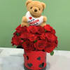 Teddy bear holding a heart pillow above red roses in a heart-decorated box