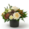Low arrangement of ivory roses and small orange flowers in a black pot with a lotus pod accent
