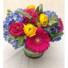 Bright bouquet of pink roses and gerbera daisy, yellow ranunculus, and blue hydrangeas in a glass vase