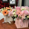 Two pastel flower arrangements in branded gift boxes with ribbon and chain handles.