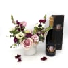 Pink and white floral arrangement in a white vase with a bottle of sparkling wine and gift box.