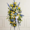 Standing floral spray with white lilies, blue flowers, and yellow roses on a white ribboned easel