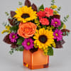 Mixed bouquet with sunflowers, roses, and daisies in an orange glass vase