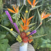 Tropical arrangement with orange bird-of-paradise and purple accents in a white vase