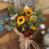Rustic bouquet of yellow sunflowers, orange roses, and burgundy blooms wrapped in brown paper and raffia.