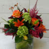 Orange roses and lilies in a clear square vase with green and burgundy accents