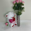 Pink roses in a tall silver vase beside a white teddy bear holding hearts