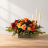 Low floral centerpiece with orange, red, and yellow roses beside three taper candles