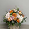 Round arrangement of peach and orange roses, hydrangeas, and pink blooms in a woven basket