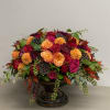 Low arrangement of orange and red roses with dark accent flowers in a bronze pedestal bowl
