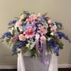 Large pastel floral arrangement with pink lilies, roses, and blue flowers on a draped stand