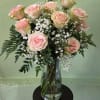 Tall arrangement of light pink roses and white filler flowers in a clear glass vase on a small dark table.