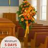 Standing spray of bright orange daisies with yellow bow on an easel beside a wood casket in a chapel.