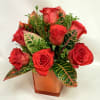 Red roses arranged in an orange square vase with variegated leaves