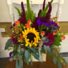 Colorful mixed bouquet with a sunflower, orange roses, and purple blooms in a rustic container.