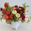 Autumn arrangement with red and orange flowers, mini pumpkins, and an apple in a white ceramic vase