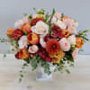 Rounded arrangement of pink roses, orange tulips, and mixed blooms in a white pedestal vase.