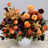Low white vase filled with orange and burgundy flowers including roses, orchids, dahlias, and calla lilies