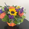 Bouquet with a sunflower, coral roses, and purple flowers in an orange pumpkin container
