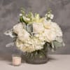White roses and hydrangeas in a glass vase with a small candle beside it