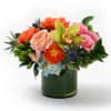 Low arrangement of orange and pink roses, green orchid, and mixed blooms in a clear glass cylinder vase.