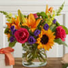 Bright mixed bouquet in a clear glass vase with roses, lilies, sunflowers, and purple blooms