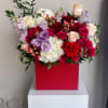 Lush arrangement of red and pink roses with white hydrangeas and purple blooms in a red square box