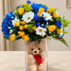 Bouquet of yellow roses, blue carnations, and white daisies in a glass vase with a teddy bear