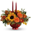 Autumn floral arrangement with sunflowers, roses, and two red taper candles in a red bowl